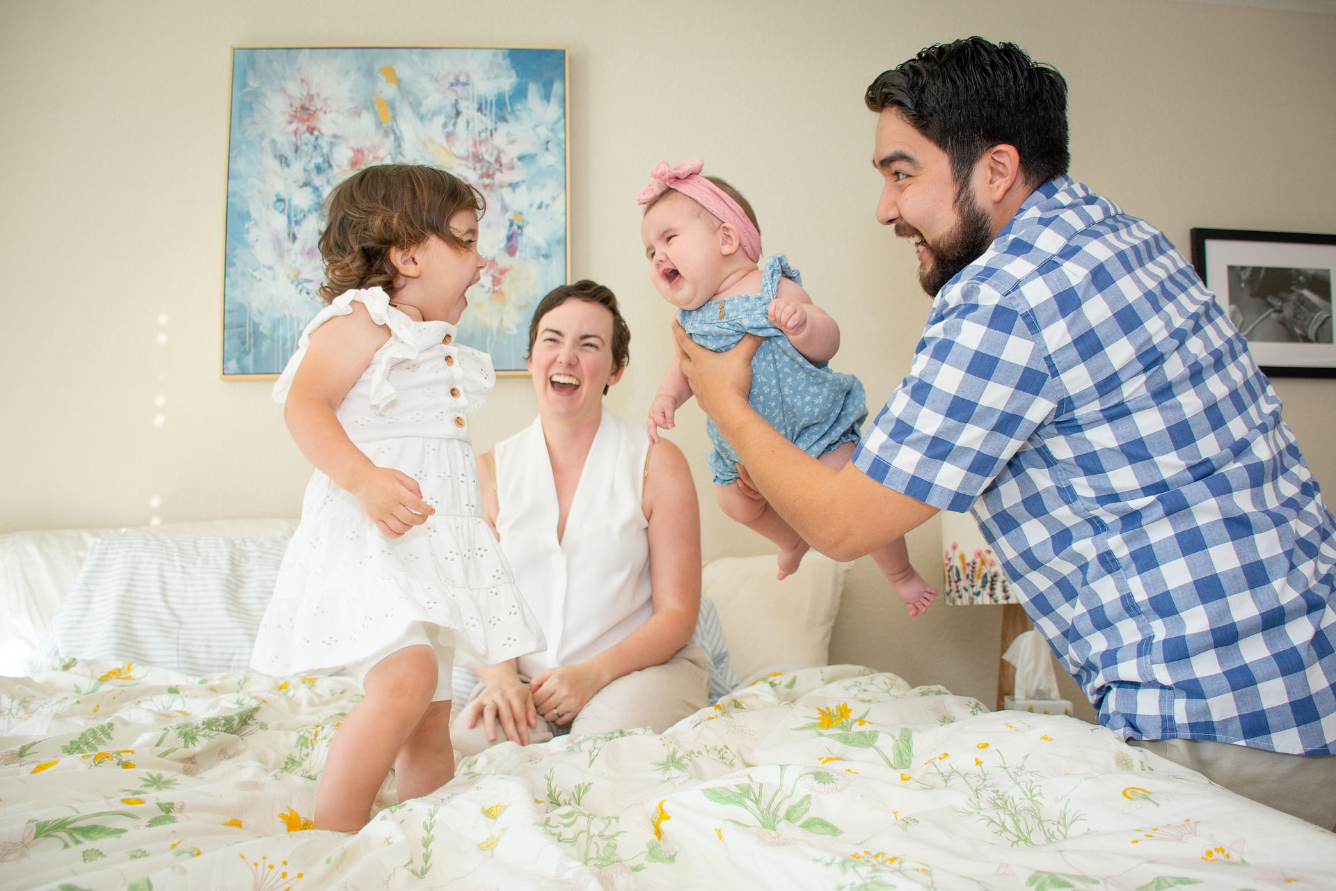 A lifestyle family picture of a father holds his baby daughter and lifts her toward her sister to make her laugh.