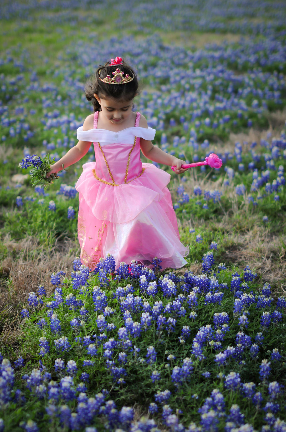 A girl wearing a pink dress and a princess crown walks through bluebonnets.
