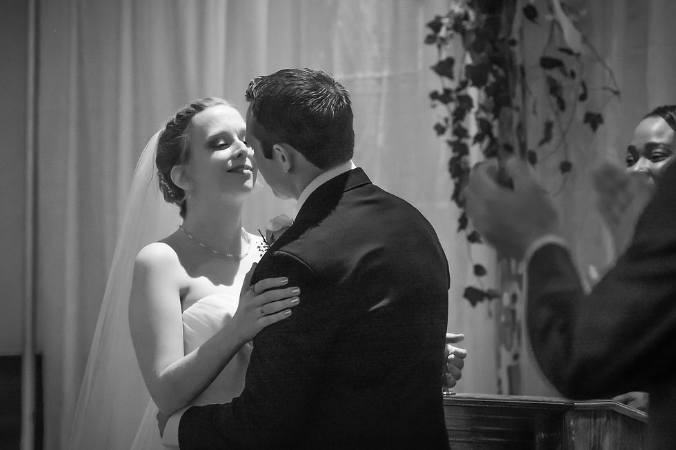 Bride and groom share a kiss during their wedding ceremony, surrounded by clapping guests. Elegant setting with floral decor.