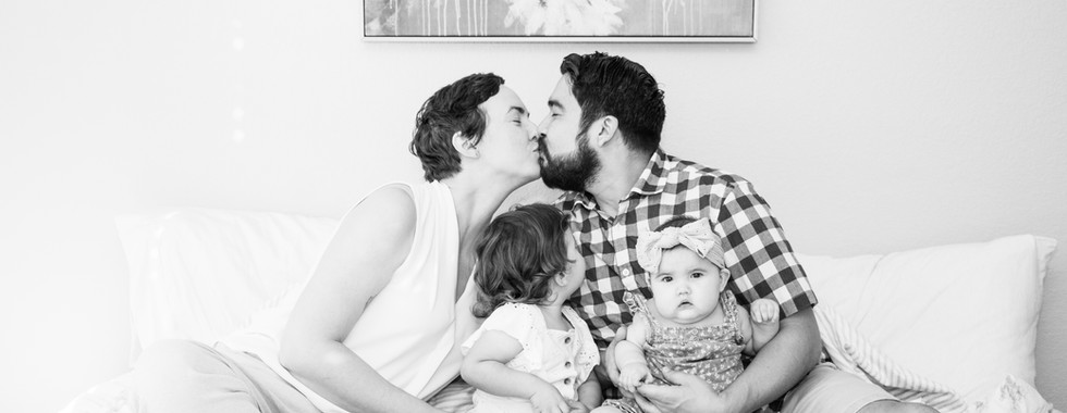 A black and white photograph of a mother and father kiss holding their 2 daughters on top of their bed.