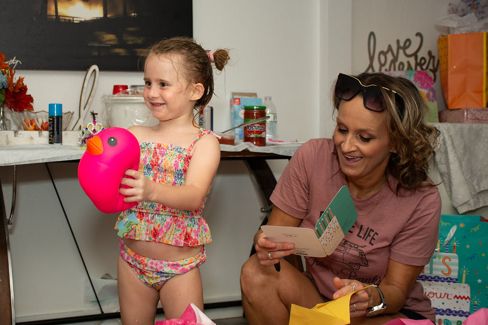 Young girl in floral swimsuit holds a pink toy. Woman in pink shirt reads card, surrounded by gifts. Decor says "love" in background. Smiling.