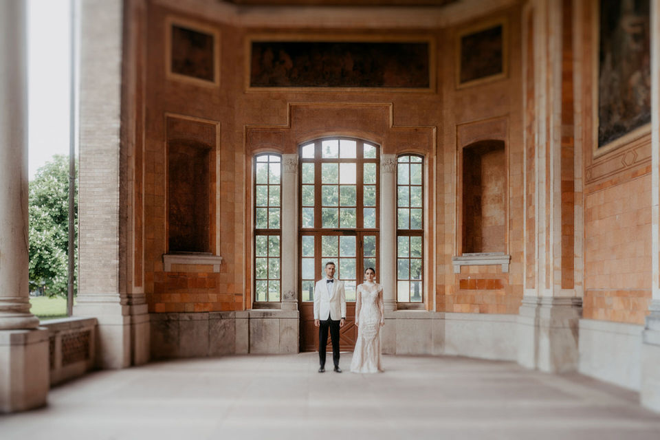 Braut und Bräutigam stehen eng beieinander in einer historischen Säulenhalle und blicken ernst in die Kamera.