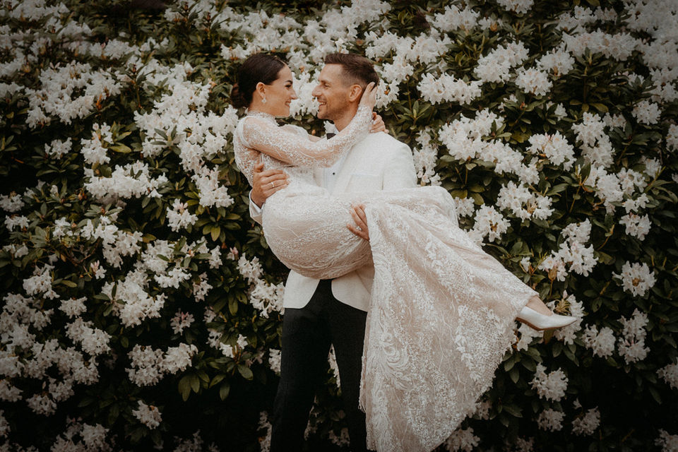 Porträt der Braut vor üppigen weißen Blüten, elegant und ruhig in ihrem Spitzenkleid.