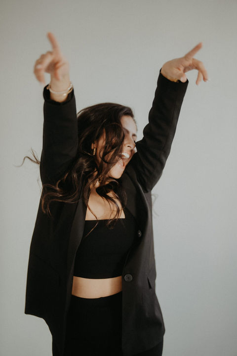 Frau tanzt mit erhobenen Armen in schwarzer Jacke im hellen Studio.