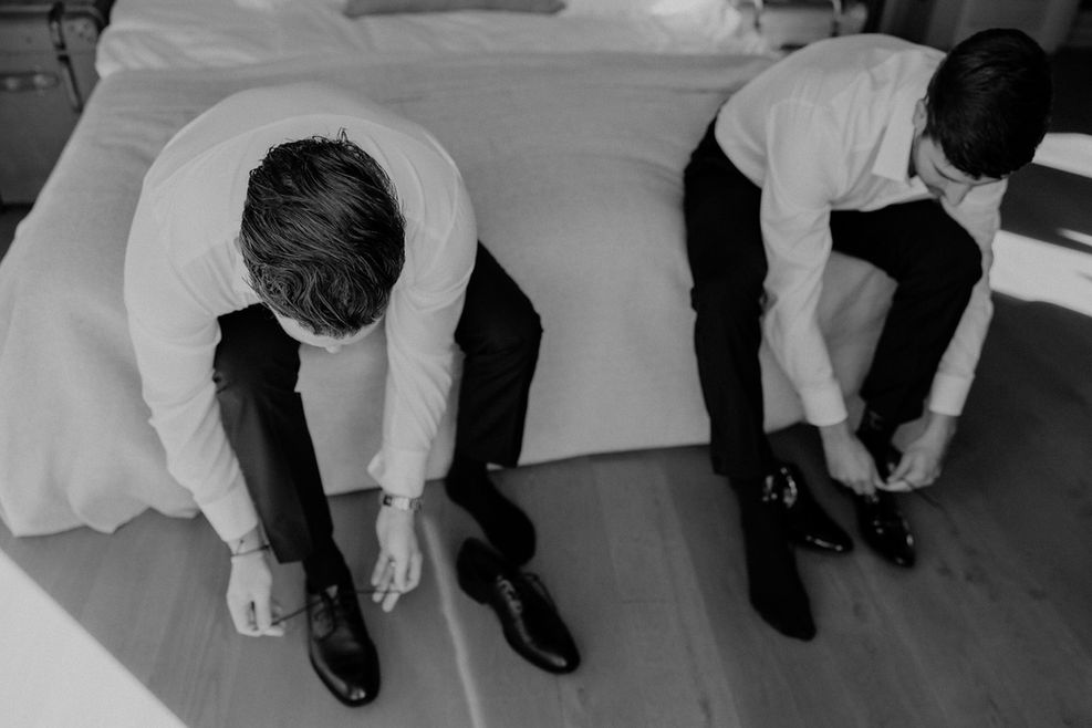 Zwei Groomsmen binden ihre Schuhe im Hotelzimmer, dokumentarischer Moment des entspannten Getting Ready.