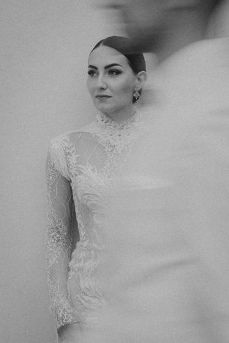 Bräutigam lehnt lässig an einer weißen Wand, während die Braut im eleganten Kleid bewegt im Vordergrund vorbeigeht.