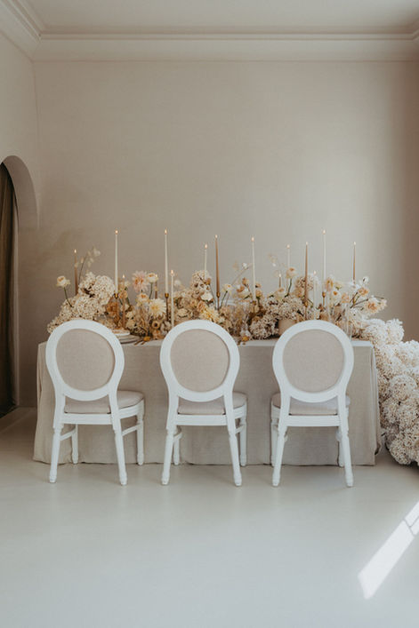 Edle Hochzeits-Tischdekoration mit cremefarbenen Blumen, hohen Stabkerzen und stilvollen weißen Stühlen. Luxuriöses, modernes Setup für elegante Events und Weddings.