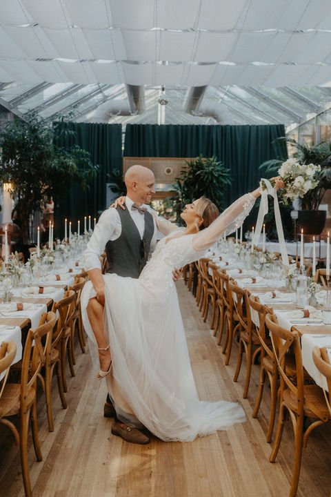 Braut und Bräutigam tanzen zwischen langen Dinner Tables im festlich dekorierten Glashaus – eleganter, moderner Wedding Moment.