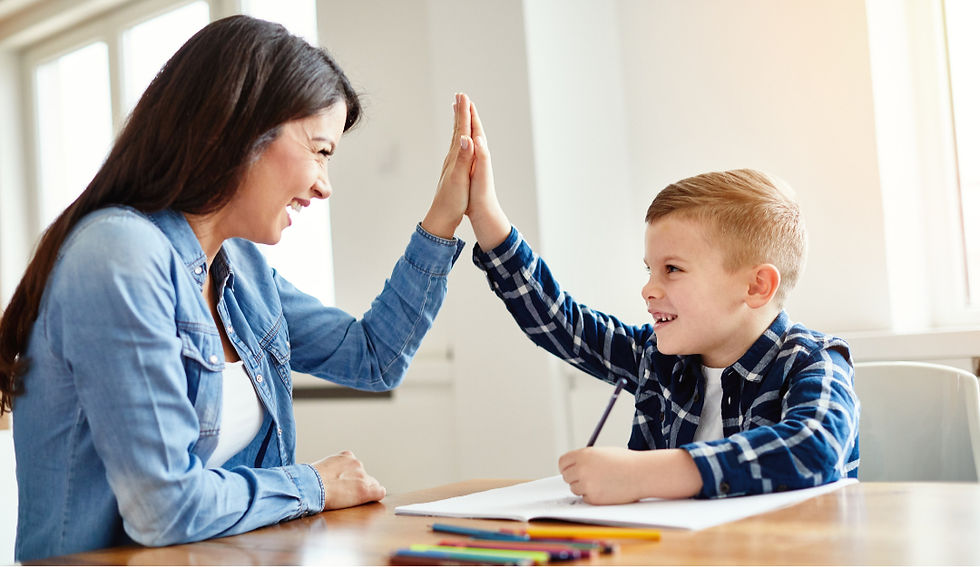 A woman and child share a joyful high-five at a table. The child holds a pencil, with papers and colored pencils on the table. Bright setting.