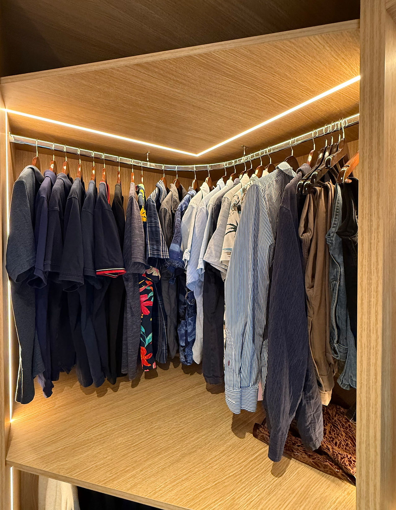 Lit wooden corner closet featuring various clothes on hanging racks.