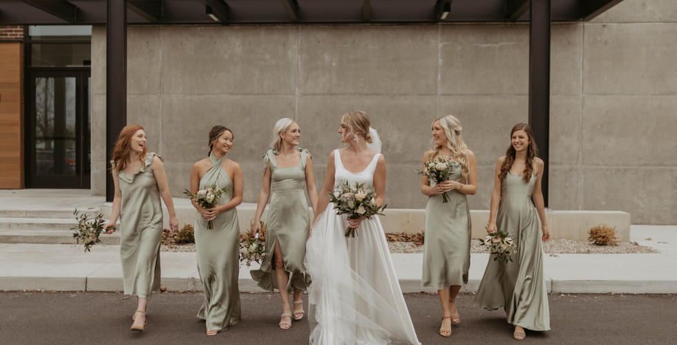 beautiful bridal party walking in front of their michigan wedding venue, Leona Road in Grand Rapids Michigan. The girls are dressed in satin sage dresses of all different styles. They laugh as they are taking photos, just before their indoor ceremony.