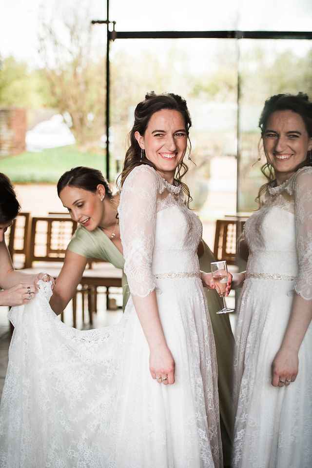 Bride in wedding dress being helped by bridesmaids with reflection in glass. Gay wedding photography at Dodford Manor wedding venue for key workers. Natural wedding photography. East Midlands Wedding Photography. Covering wedding photography in Staffordshire, Derbyshire, Northamptonshire, Lincolnshire, Darley and Underwood Photography.