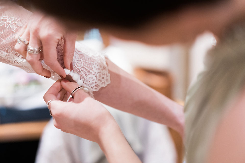 Detail wedding photograph of bride's lace sleeve on her wedding dress. Bridesmaid helping in olive dress. Wedding photography, Northamptonshire.