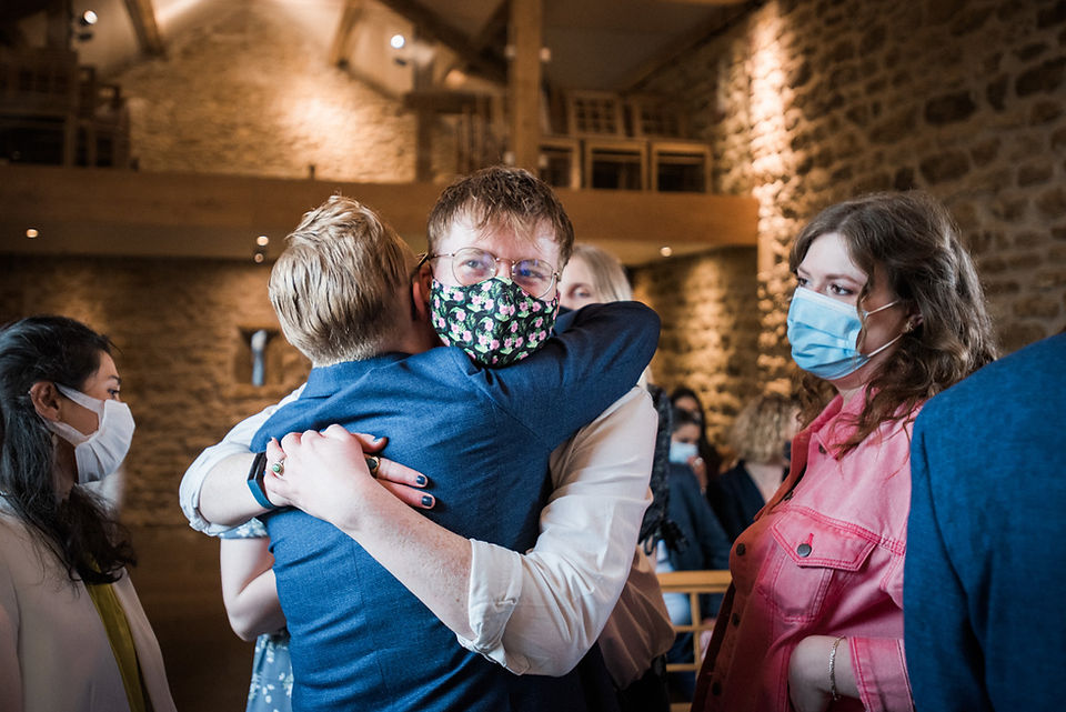 Wedding guest hugs bride in suit. Guest wearing colourful wedding face mask at Dodford Manor. Candid wedding photography in Northamptonshire.