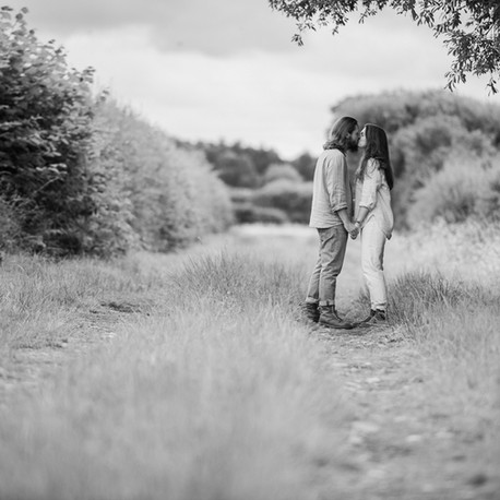 Engagement photograph. A black and white landscape photograph of a couple kissing on a country lane, surrounded by fields. Shot by Darley and Underwood photography, specialist in wedding photography on both digital and film. Shot in York, North Yorkshire