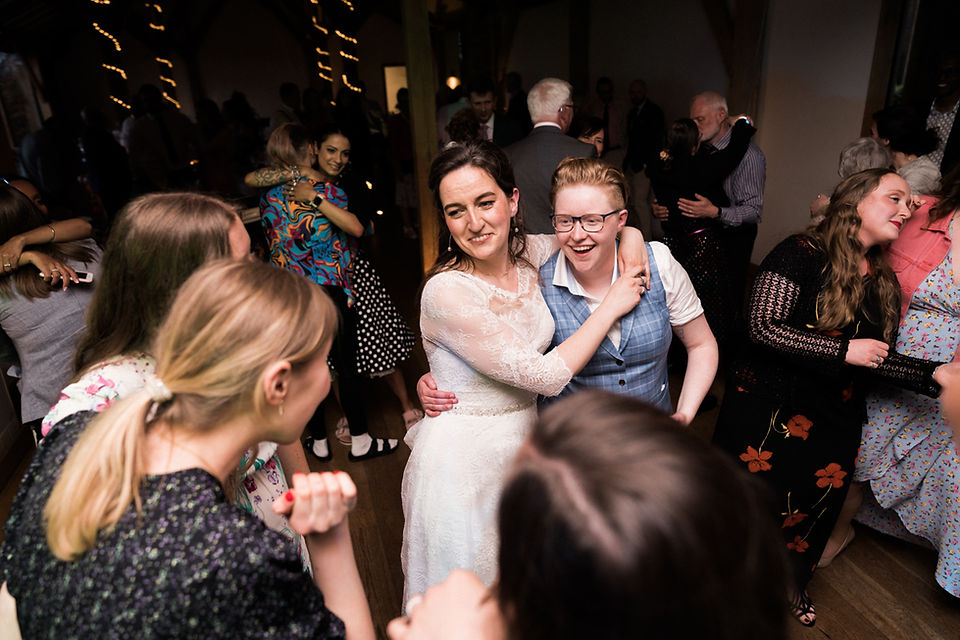 Bride and bride couple embrace on the dancefloor with a crowd of wedding guests. Smiling and laughter. 
