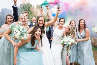 Gay wedding photography Leicestershire. Bride and bride with wedding party huddled and laughing with wedding smoke bombs / grenades in the air. Fun, relaxed wedding photography in Leicestershire. Same sex couple both in white wedding dresses surrounded by bridesmaids in light blue bridesmaids dresses and wedding bouquets everywhere. Fun filled wedding chaos. 