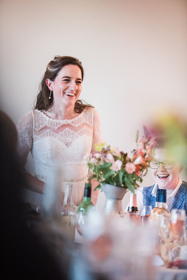 Bride in lace wedding dress making her new bride laugh during wedding speech. Gay wedding photography at Dodford Manor, Northamptonshire. Relaxed wedding photography. Leicestershire wedding photography. Covering wedding photography in Warwickshire, Derbyshire, Northamptonshire, Darley and Underwood Photography.