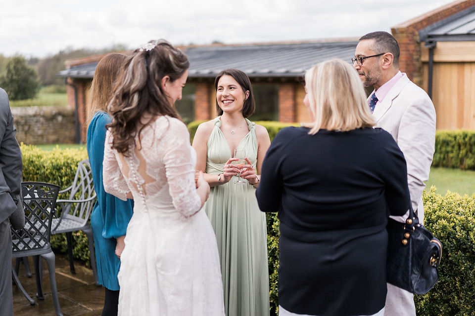 Fun wedding photography capturing guests having a laugh in the courtyard at Dodford Manor, Northamptonshire. Bridesmaid laughing in olive dress in group of five wedding guests. Natural wedding photography. Leicestershire wedding photographer. Covering wedding photography in Warwickshire, Staffordshire, Derbyshire, Lincolnshire, Nottinghamshire. Darley and Underwood Photography.