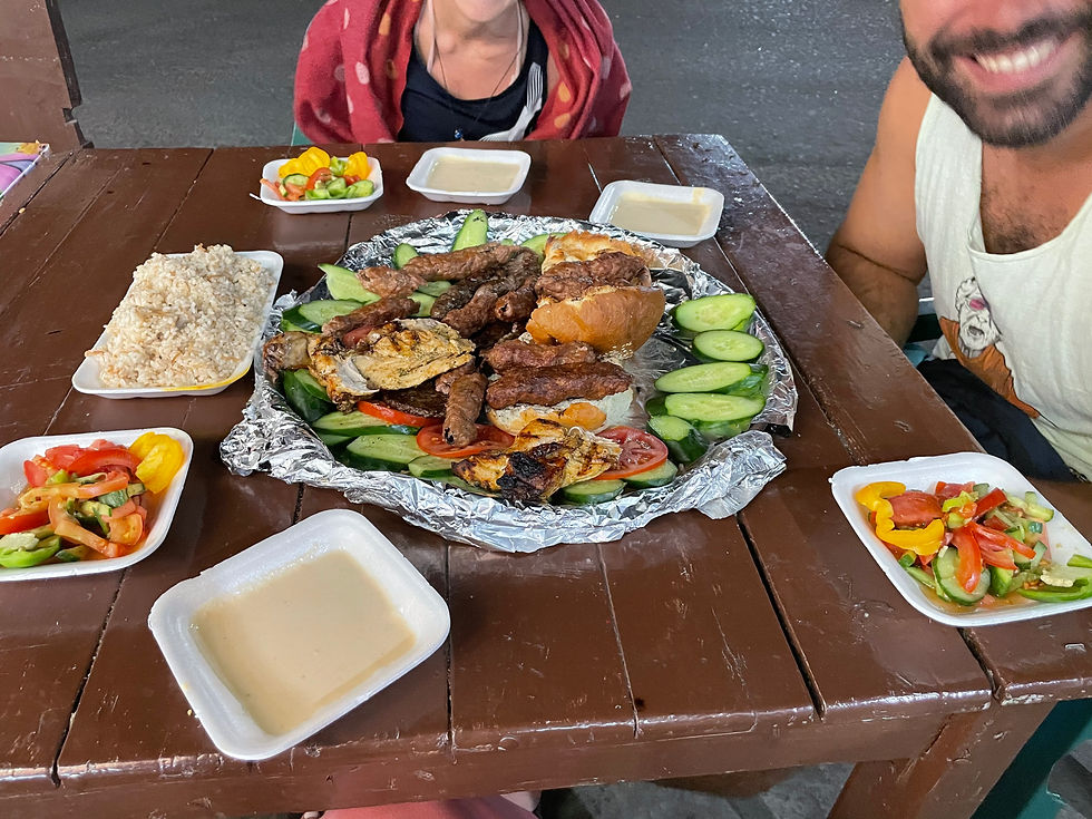 Platter of grilled meats and vegetables on foil, surrounded by salad, rice, and dips on a wooden table. Two people dining outdoors.