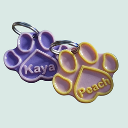 Twee 3D-geprinte hondenpenningen in pootvorm met de namen Kaya en Peach in pastel paars en geel.