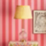Vintage lamp with yellow pleated shade on floral tablecloth. Pink striped wallpaper and framed artwork in background. Vibrant and retro.
