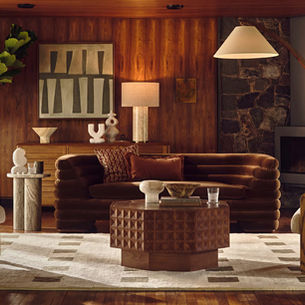 Cozy living room with brown sofa, patterned rug, wooden furniture, plants, and abstract art. Warm lighting and a stone fireplace create a tranquil vibe.