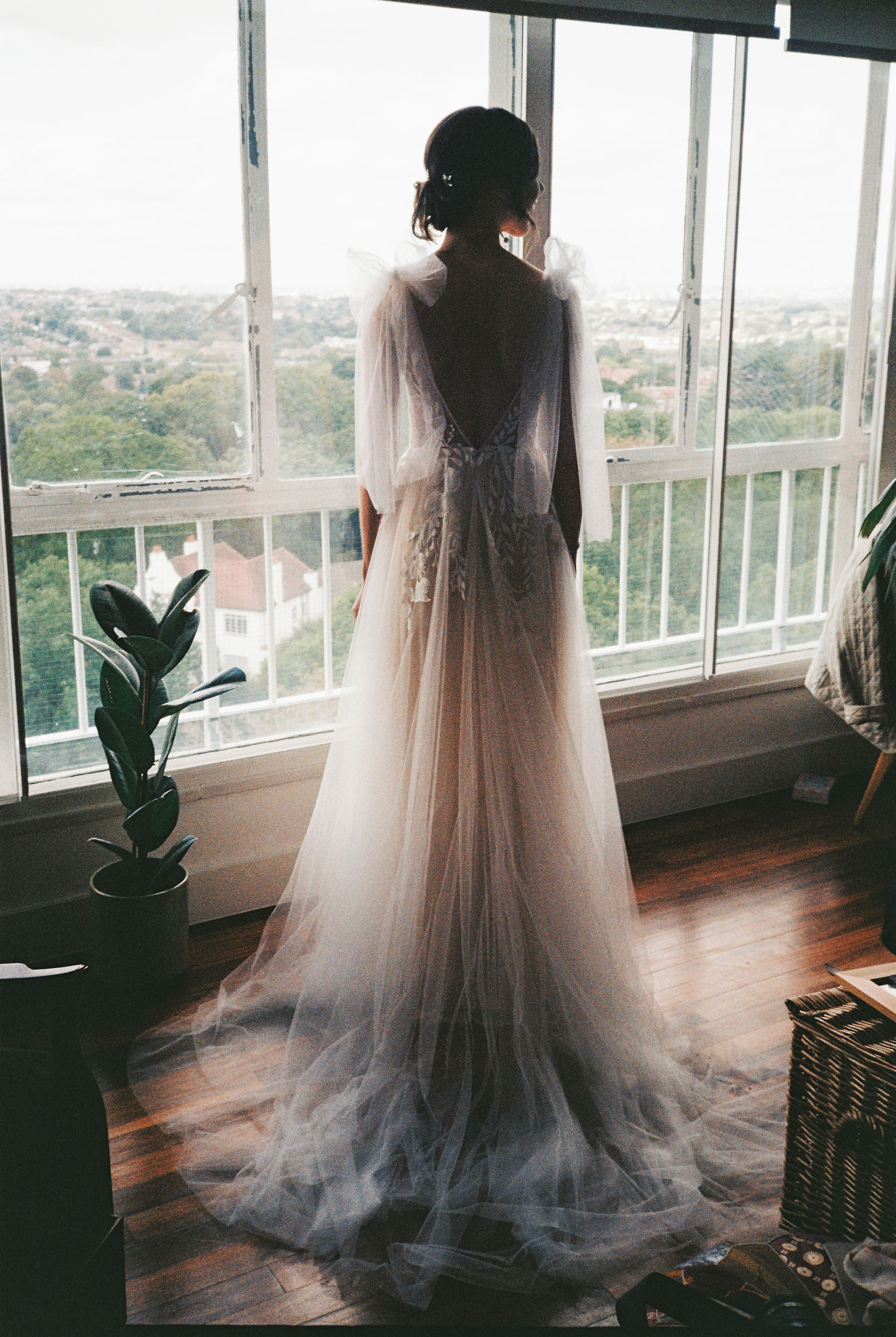 Bride is standing in front of a window over looking London. Her gown highlighted by the day light. Boho Bride. Image taken on 35mm Film Camera