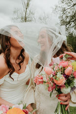 bride and bride laughing with each other as their veil has blown over their heads. Boho wedding vibes with flowers.
