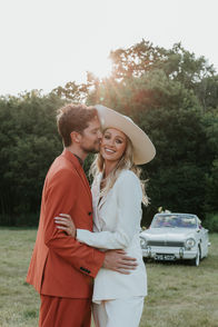 Bride and groom are both smiling. They stand in a field in Sussex and the two woods estate with a vintage car in the background. The groom is kissing her cheek and the bride is smiling to the camera. It is golden hour and the sun is beating down behind them.
Ellamae Cieslik photography