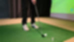 Close-up of a golfer in dress shoes addressing a golf ball on indoor turf next to a simulator screen.