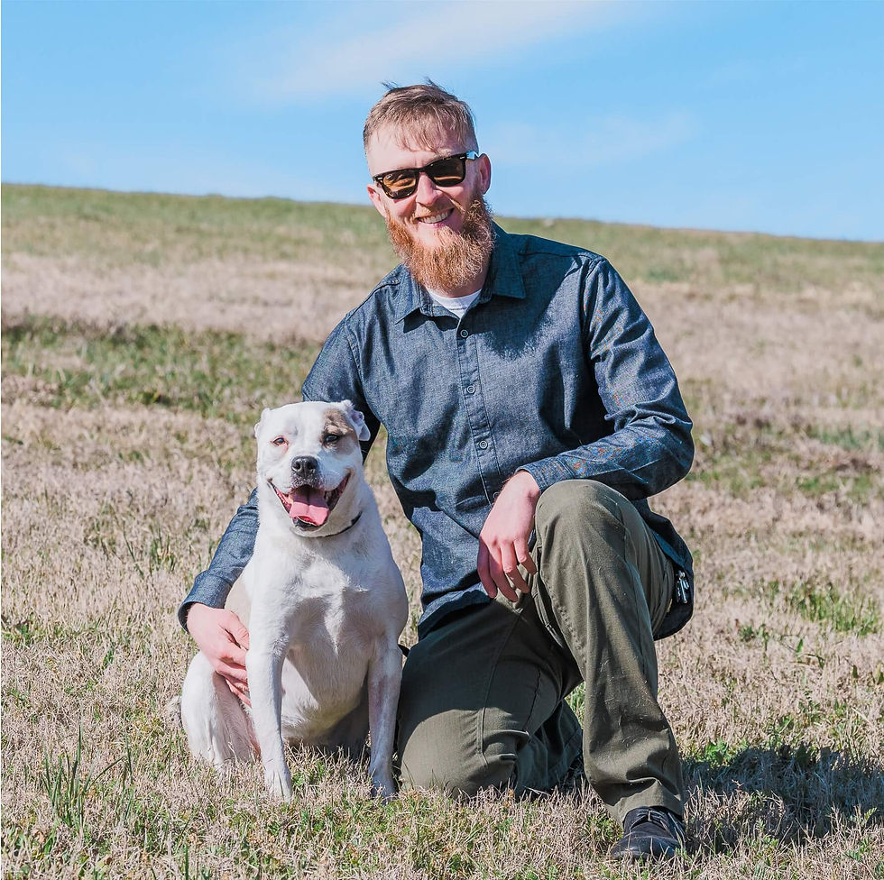 A man with a beard and sunglasses kneels beside a smiling white dog on a grassy hill under a clear blue sky