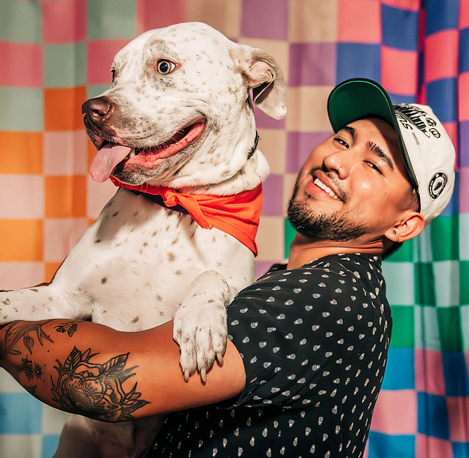 A man joyfully holds a large, spotted dog wearing a red bandana. They are in front of a colorful checkered background.