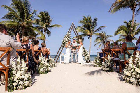 Two brides exchanging vows under a triangle arch with palm trees swaying in the background.