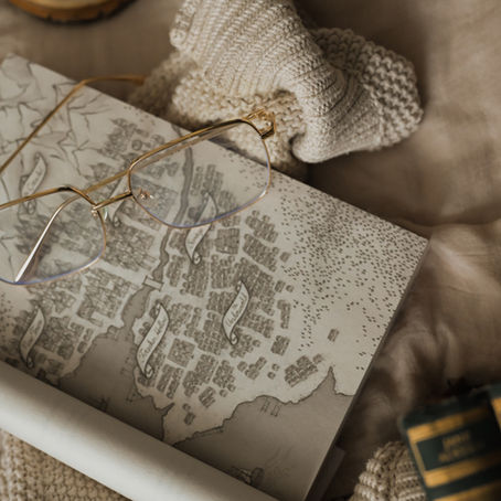 Gold-rimmed glasses on an open map book beside a cream knitted scarf and candle, in a cozy beige-toned scene