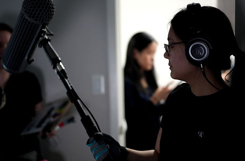 Boom op Kathy Tu recording audio on set - set is a bedroom but in this image it is not identifiable as a bedroom. only a grey wall and a light switch is visible 