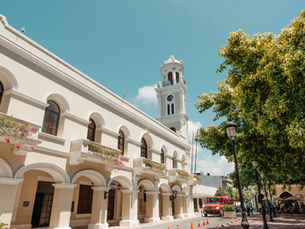 A white building with arches and a clock tower beside leafy trees in Santo Domingo, Dominican Republic.