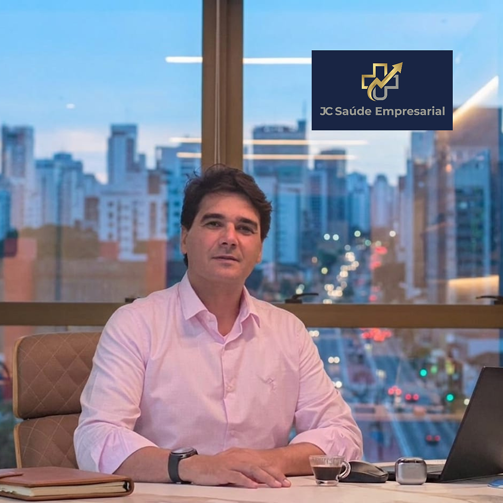 João Coelho, CEO e Co-Founder da JC Saúde Empresarial sentado em uma mesa de escritório com vista urbana ao entardecer ao fundo, diante de notebook e logo JC Saúde Empresarial.