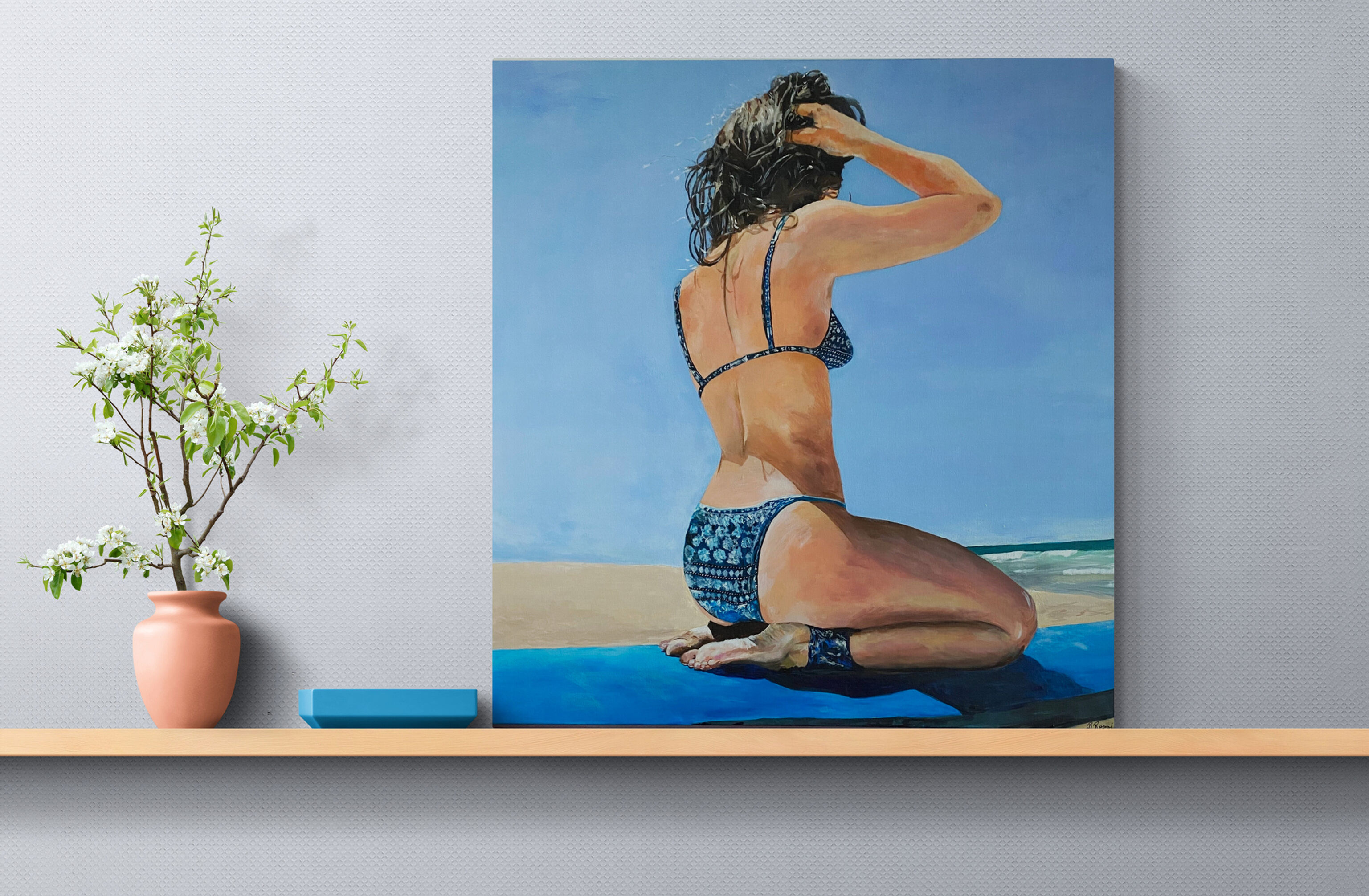 Young woman in a blue patterned bikini sits cross-legged on a surfboard near the shoreline, one hand raised to adjust her hai