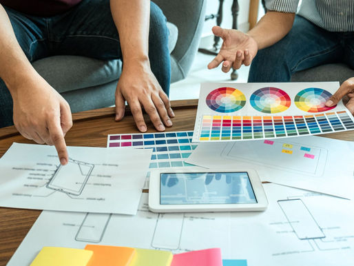 Two people discuss design plans on a table, pointing at color swatches and sketches. A tablet and colorful papers are present.