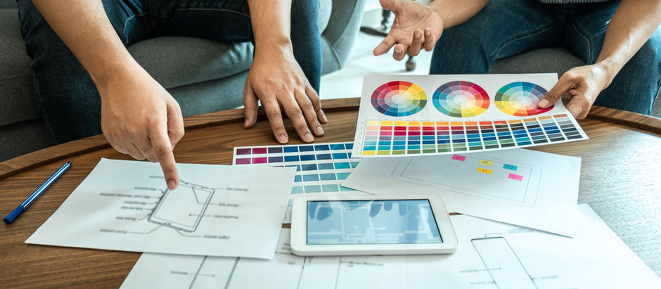 Two people discuss design plans on a table, pointing at color swatches and sketches. A tablet and colorful papers are present.