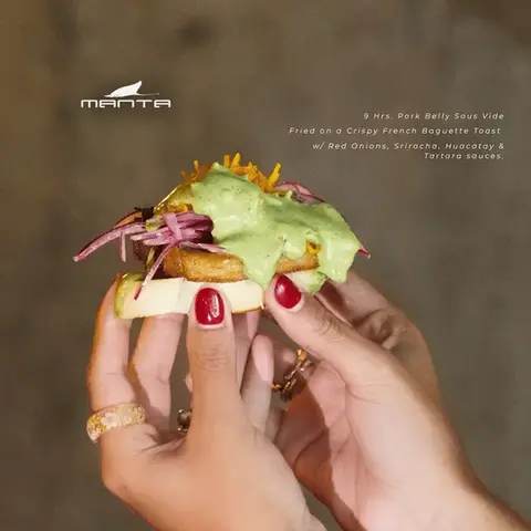 Hands holding a Manita New York Deli Style sandwich with lettuce, sprouts, and crispy filling on a French baguette.