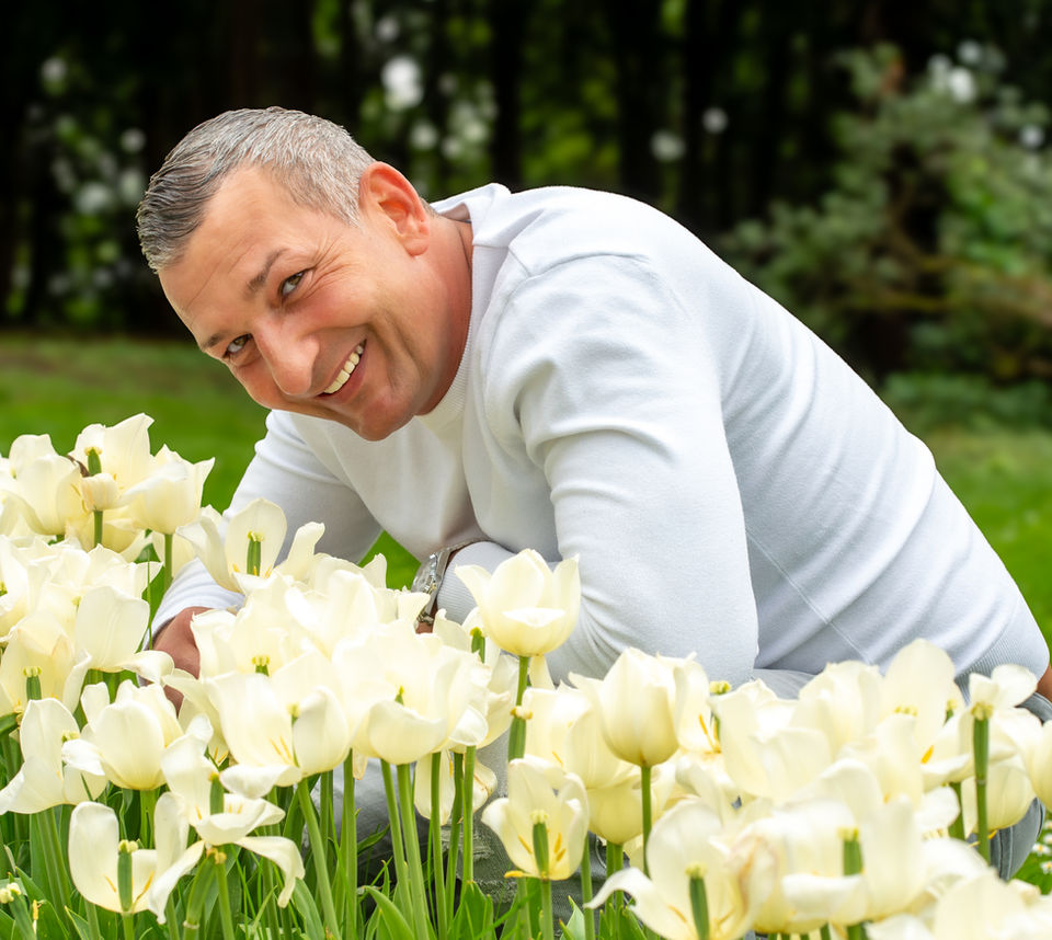 Independent Escort Kevin für Frauen und Paare hockt vor Blumen und lächelt in die Kamera