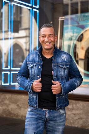 Callboy Kevin trägt ein schwarzes Shirt dazu eine blaue Jeans Jacke sowie eine blaue Jeans Hose und lächelt in die Kamera