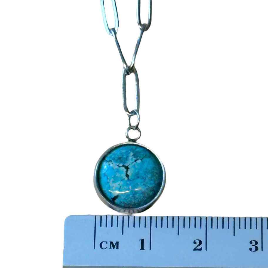 A close up of a round blue turquoise charm against a ruler.