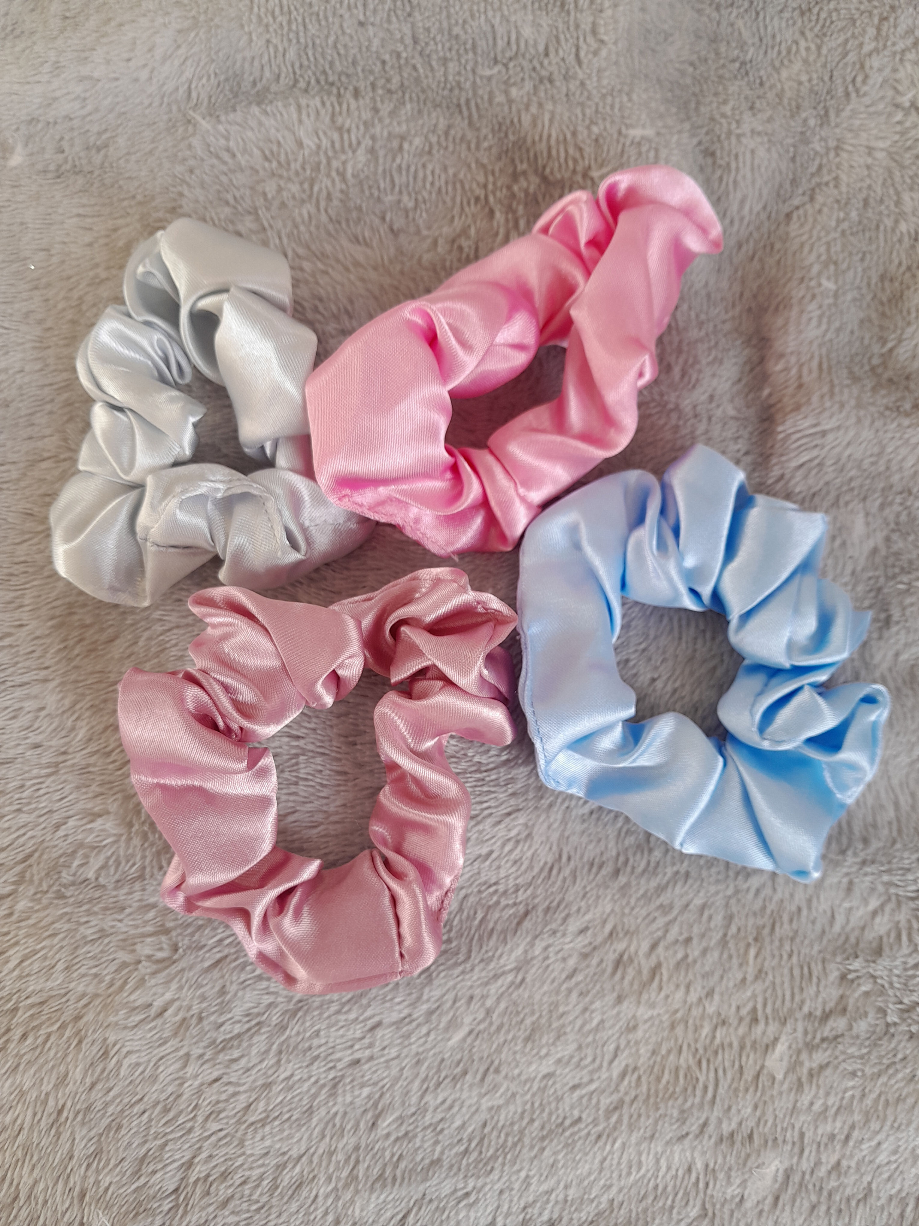 Quatre chouchous de satin rose, bleu clair et gris, posés sur tissu.