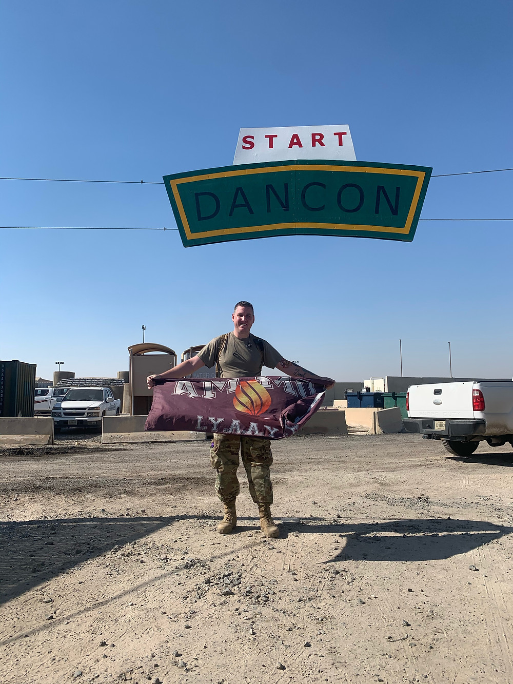Derek holds a flag that says "Ammo" under a banner that says "Start Dancon". There are cars around him. He seems to be in a desert parking lot.