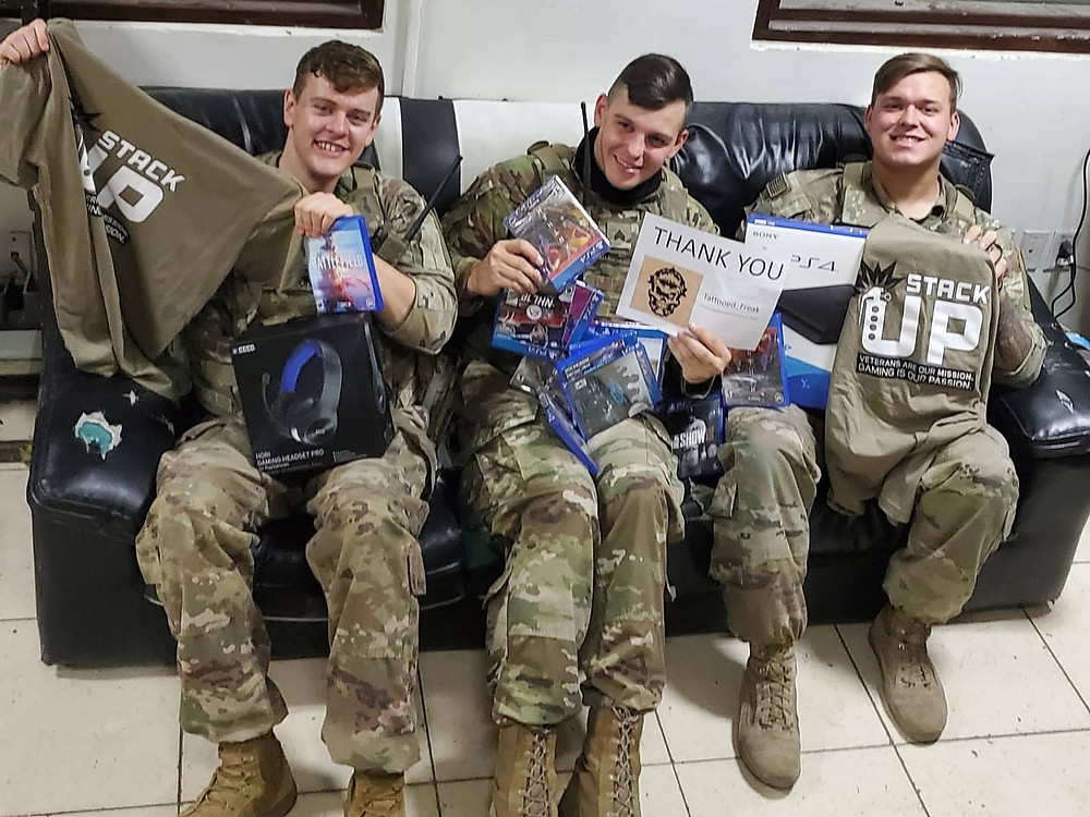 3 soldiers sit on a couch. They are holding up PlayStation 4 games, and stack up t shirts.