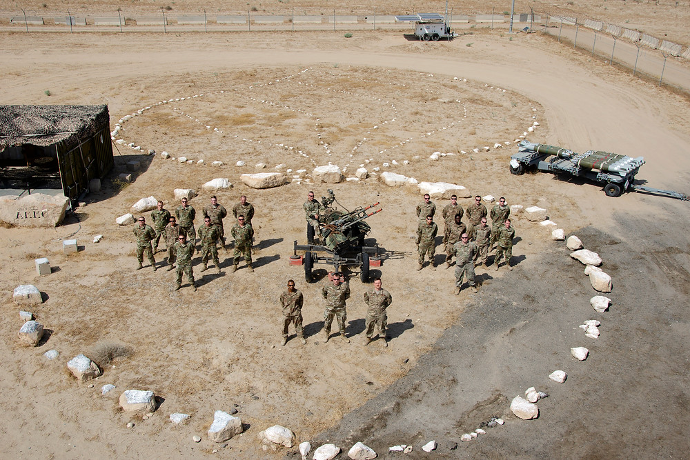 Derek's unit stands in formation in Army uniform inside of a circle made of white rocks. They are in the desert.