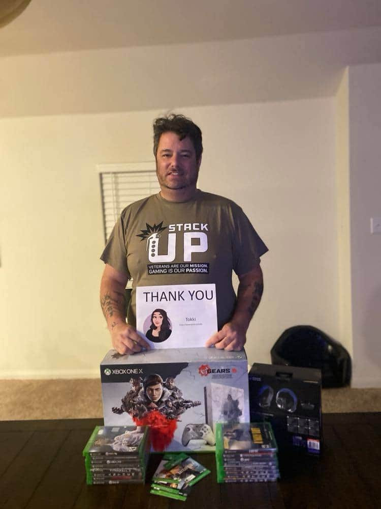 Giles stands behind his new Xbox One X. He holds a thank you note up for Tokki the sponsor of his crate. He has stacks of videogames in front of the Xbox.